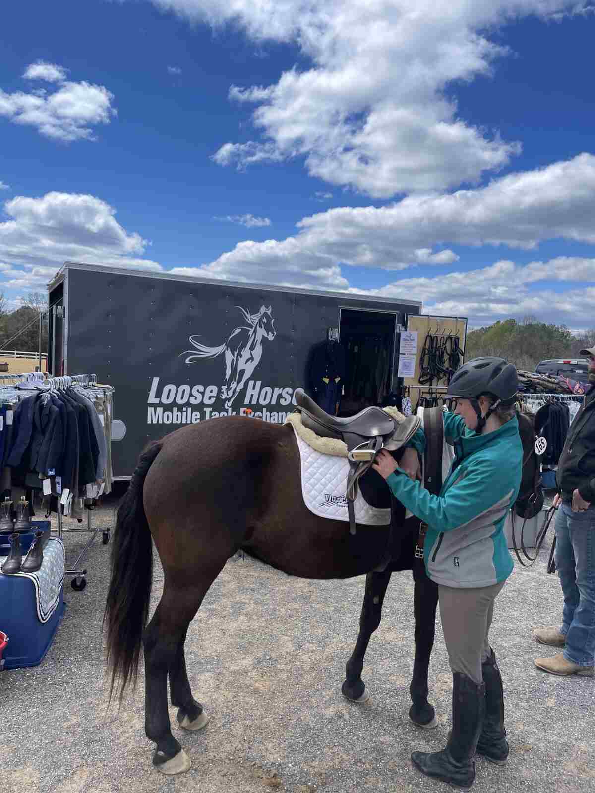girl in a green jacket with riding helmet tacking up a bay horse by loose horse trailer